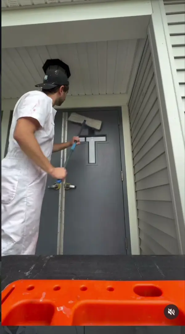 Painter in white coveralls sanding the top of a gray exterior door under a porch